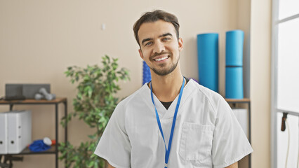 Portrait of a smiling young hispanic male healthcare professional wearing a lab coat with a lanyard in a rehab clinic.