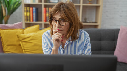 Middle-aged woman with glasses and short hair, sitting thoughtfully in a living room on a sofa, watching television indoors.