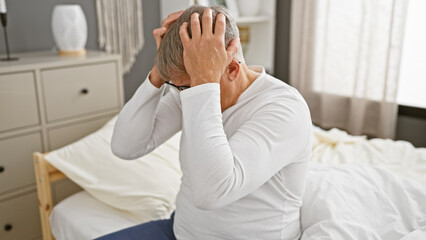 A distressed grey-haired man sitting on a bed in a bedroom, portraying a scene of worry or headache.