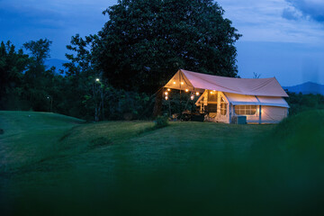 A small white tent is set up in a grassy field. The tent is lit up with lights, giving it a cozy and inviting atmosphere. The scene is peaceful and serene, with the tent providing a sense of shelter