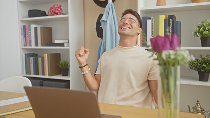 Joyful hispanic man celebrating success indoors at a home office with books and plants around him.