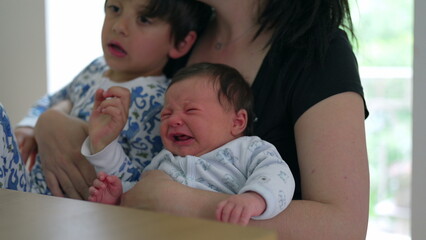 Mother holding her baby and young child, comforting a crying baby. family is sitting at a table, showcasing the challenges and love in parenthood. Children are in cozy pajamas