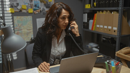 A middle-aged woman detective is working at her desk in an office, talking on the phone and using a...
