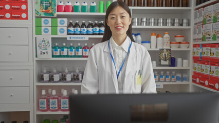 A young asian female pharmacist smiling in a modern pharmacy interior surrounded by medicine shelves.