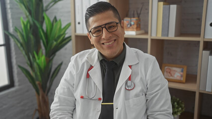 A smiling hispanic man in a white lab coat with a stethoscope poses confidently in a clinic office.