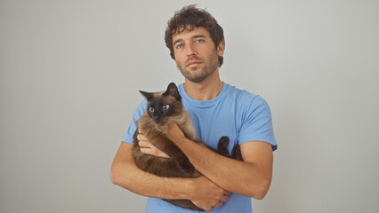 Obraz premium A young hispanic man in a blue shirt holding a siamese cat against an isolated white background.