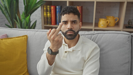 A young hispanic man gestures disapprovingly with his middle finger while sitting in a cozy home living room.