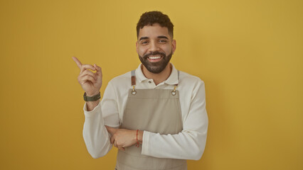 Handsome hispanic man smiling and pointing in apron over yellow background