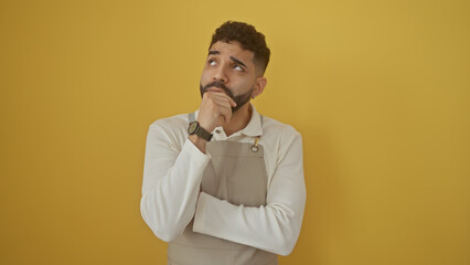 Pensive hispanic man with beard wearing apron over isolated yellow background, thinking.