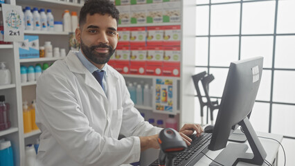 Handsome hispanic pharmacist works at computer in modern drugstore