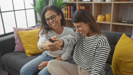 Two women, likely mother and daughter, cheerfully struggle for a remote control in a cozy living room.