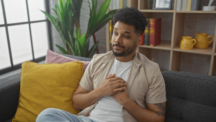 Handsome black man with hands clasped on chest sitting thoughtfully in a cozy living room.