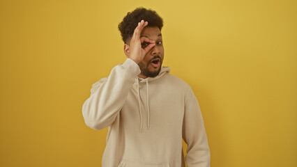 African american man making ok gesture over eye against a yellow wall