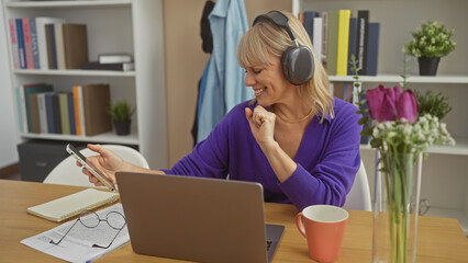 A cheerful woman in a purple sweater enjoys music on her headphones while using a smartphone in a cozy living room with a laptop and flowers on the table.