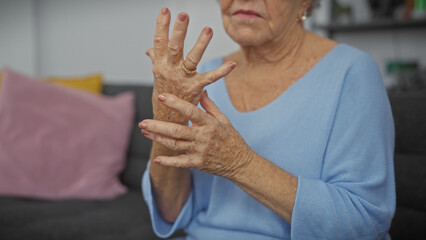 A senior woman with grey hair examines her hands for signs of aging or health issues in a cozy living room.