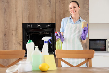 Young woman with sponge and different bottles of detergent in kitchen