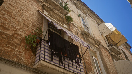 Rustic balcony in bari, italy, with striped awning and clothes drying, framed by weathered walls and vibrant blue sky.