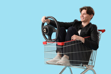 Cool young man with steering wheel in shopping cart on blue background