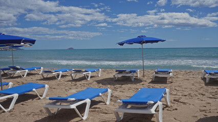 Blue sunbeds and umbrellas on a sandy beach overlooking the calm sea under a cloudy sky, depicting a typical summer vacation scene.