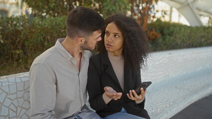 A man and woman discuss seriously while seated on a park bench with a smartphone in her hand, portraying a relationship in an urban outdoor setting.