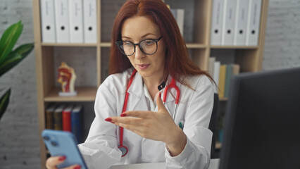 A professional female doctor in glasses conducts a telehealth consultation from her office using a smartphone.