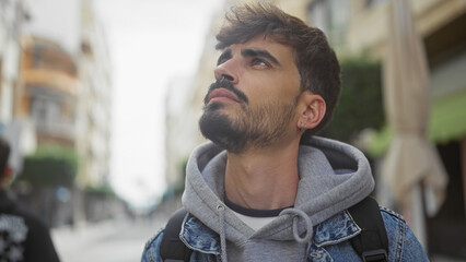 A young hispanic man with a beard gazes contemplatively while standing on an urban street outdoors.