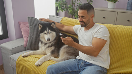 A handsome hispanic young man with a remote control sitting on a yellow couch indoors with his husky dog in a cozy living room.