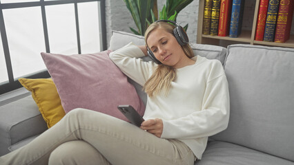 Obraz premium A relaxed young woman enjoying music with headphones and smartphone in a cozy living room on a sofa