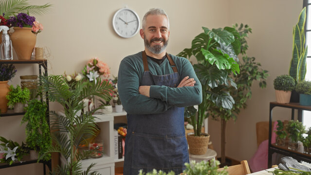 Bearded man in apron smiling confidently at a vibrant indoor flower shop with lush green plants