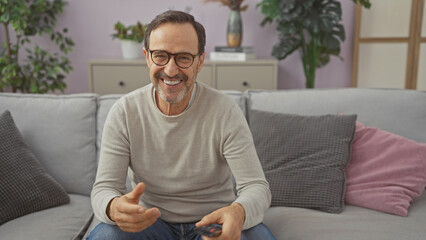 Fototapeta premium Hispanic mature man smiling sitting on a grey couch in a cozy living room holding a remote