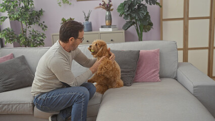 A middle-aged man affectionately pets a poodle on a sofa in a cozy living room setting, creating a homely scene.