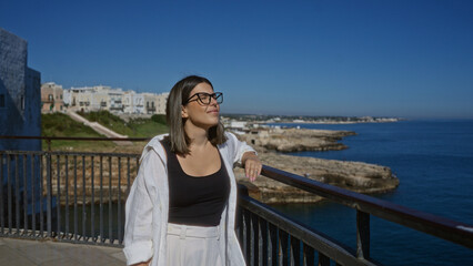 Young hispanic woman enjoying a beautiful day at a scenic viewpoint over the ocean in polignano a mare, puglia, italy, with vibrant blue skies and charming coastal buildings in the background.