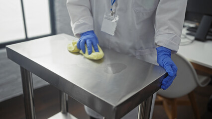 A woman wearing blue gloves cleans a stainless steel table in a clinic or hospital room with a...