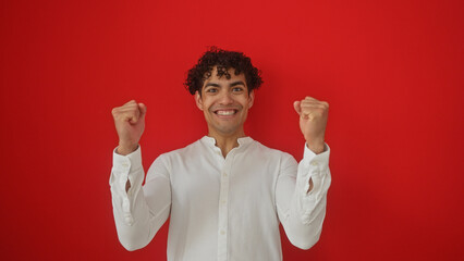 A cheerful young hispanic man in a white shirt celebrates with fists raised against a red...