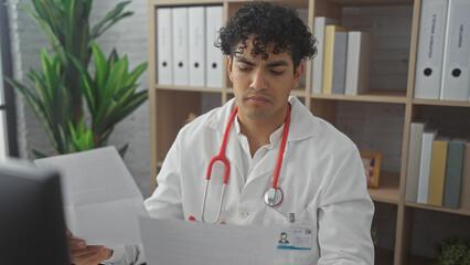 A young hispanic man in a white lab coat with a stethoscope reviews documents in a clinic office setting.