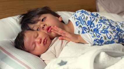 Older brother gently touches the face of his newborn sibling while both sleep, lying together on a cozy bed with soft blankets, sharing a peaceful nap time