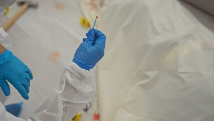 A forensic investigator collects evidence at a crime scene wearing protective gloves and a white...
