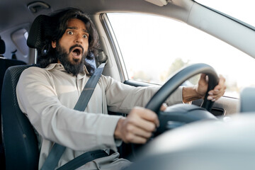 A man with long hair and a beard shows an expression of shock while driving. He clutches the steering wheel firmly, actively engaged in the unexpected moment.