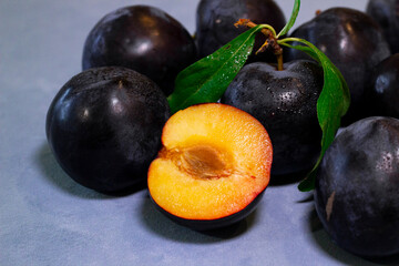 Fresh Whole and Half Black Plums with Green Leaves on Blue Backgrou Close-up of ripe black plums with one halved plum showing juicy yellow-orange flesh and pit. 