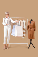 Happy mature female stylist holding shopping bags near rack with stylish clothes and mannequin on beige background