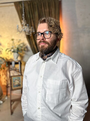 Thoughtful man in a white shirt stands confidently in a warmly lit room with a classic and elegant background interior. Confidence, elegance and male fashion concept