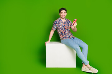 Full size photo of cheerful guy dressed print shirt jeans sit on white platform holding smartphone isolated on green color background