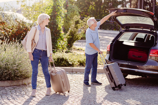 An elderly couple takes luggage out of the trunk of their car. The man holds a suitcase while closing the trunk, and the woman stands by with another suitcase, both headed towards a hotel.