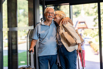 An elderly couple enters a hotel lobby, pulling their suitcases. They are dressed casually,...