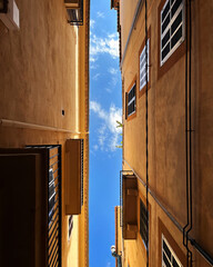 Upward view of a narrow alleyway framed by warm, sunlit buildings with a clear blue sky overhead....