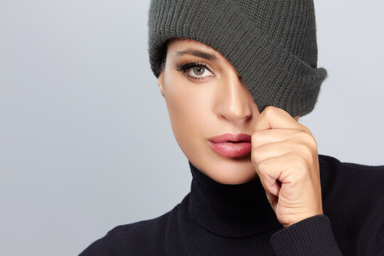 Stylish woman in black turtleneck with gray beanie covering one eye on light gray background