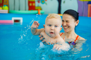 A baby learns to swim with a coach in the pool. Swimming in an early age pool. Happy child with female trainer in indoor swimming pool playing and having fun.