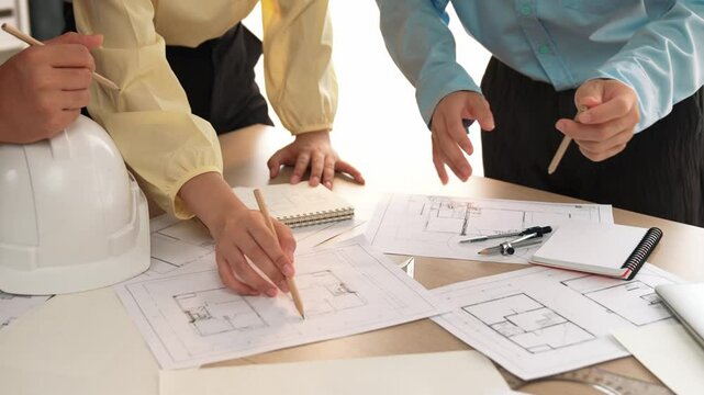 Smart architect and safety engineer discussion about building blueprint on table with architectural document, measurement tools and wooden block scatter around at modern office. Closeup. Delineation.