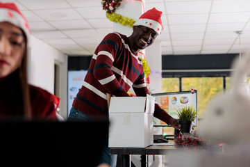 Cheerful african american employee wearing Christmas hat unpacking belongings box, decorating new desk after being hired. Businessman starting job, excited to work in festive ornate office
