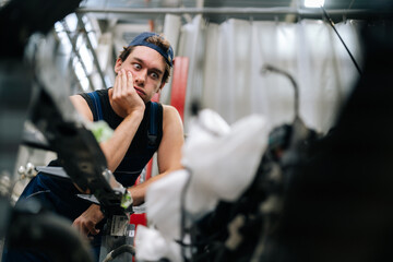 Frustrated mechanic male in uniform with thoughtful expression leaning on car part, contemplating...